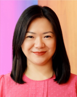 Professional headshot of a woman with straight black hair, smiling warmly and wearing a pink blouse against a colorful background.