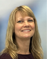 A smiling woman with straight blonde hair wearing a maroon top, posing in front of a soft blue and gray gradient background.