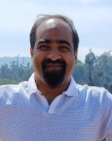 A cheerful man with a beard and mustache wearing a white and blue striped polo shirt, with trees and mountains blurred in the background.