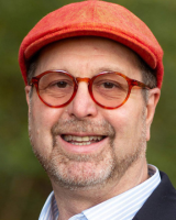 A man with a friendly smile wearing a red cap, red-rimmed glasses, and a striped shirt under a blazer.